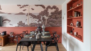 A dining area with a table set for four, a red bookshelf, and a wall mural of a landscape. - Starfish - Charmant appt à 3min à pied de la plage (Saint-Malo)
