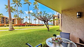 A balcony with a table set for two, overlooking a well-manicured lawn and palm trees. - Ground-floor condo with covered lanai & resort pools (Lahaina)