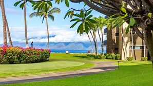 A well-maintained lawn, a curved driveway, and a building with a view of the ocean and clouds. - Ground-floor condo with covered lanai & resort pools (Lahaina)