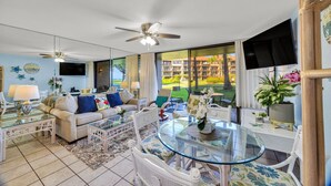 A spacious living room with a glass coffee table, white furniture, a ceiling fan, and a large window offering a view of a building and greenery. - Ground-floor condo with covered lanai & resort pools (Lahaina)