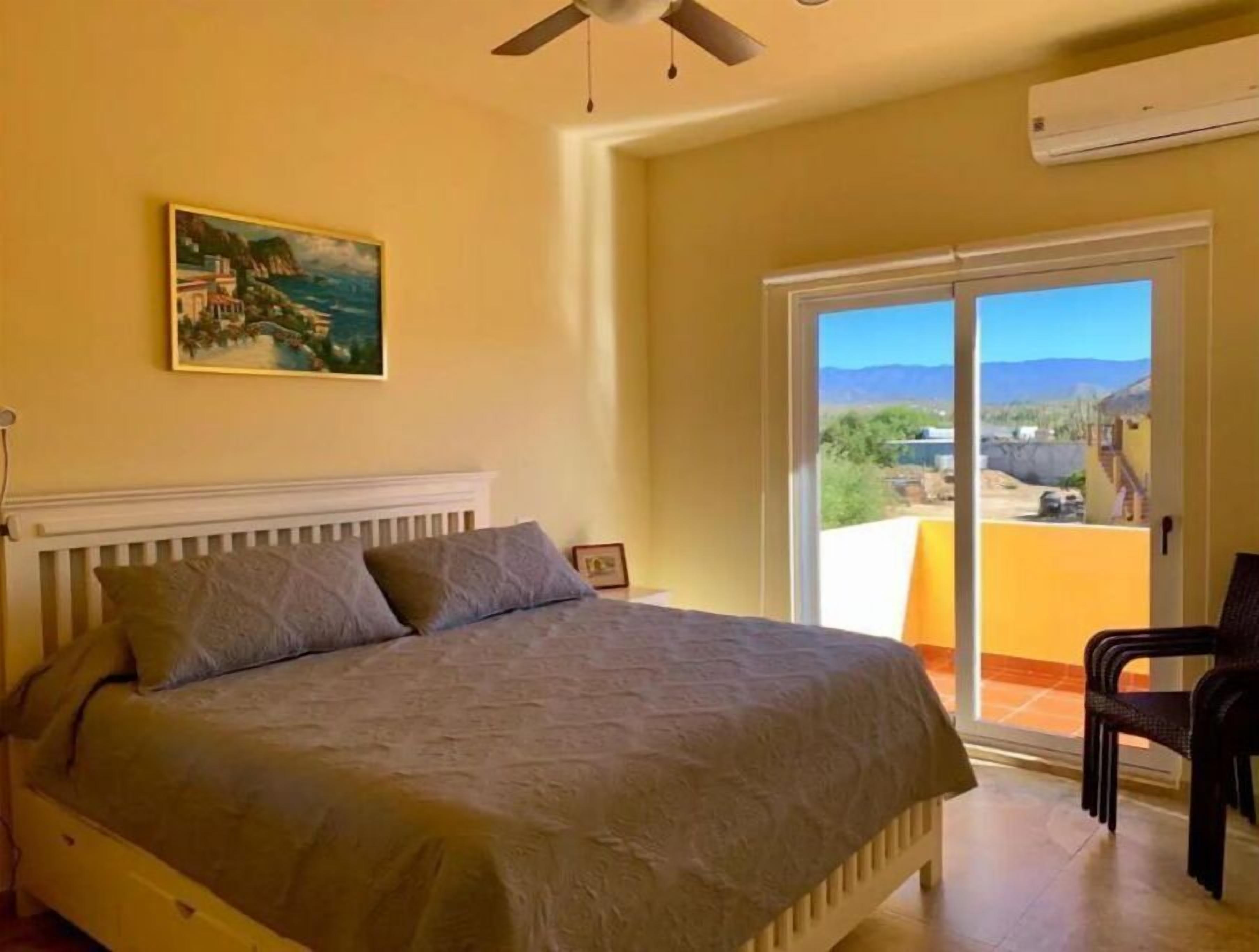 Une chambre à coucher avec un lit, un ventilateur de plafond, un tableau et une vue sur l’extérieur, visible à travers une porte-fenêtre coulissante.