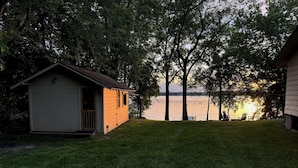 Eine kleine Holzhütte mit Veranda, einem grünen Rasen und Blick auf einen See bei Sonnenuntergang.