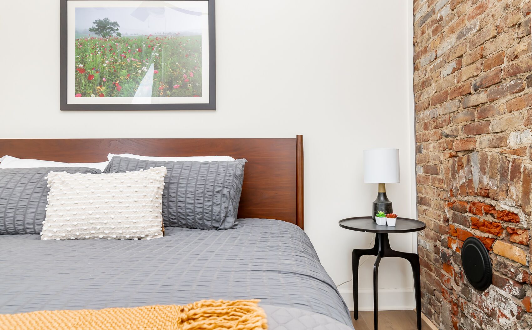 A neatly made bed with grey bedding and decorative pillows, a small round table with a lamp and two potted plants, and a brick wall.
