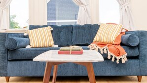 A blue sofa with striped pillows and a macrame throw, a small white table with books, and a window with curtains.