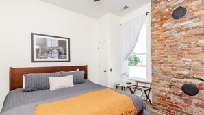A bedroom with a bed, a framed picture on the wall, a brick wall, and a window with curtains.