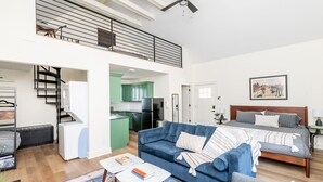 A spacious living area with a blue sofa, a bed with a wooden headboard, and a kitchen with green cabinets. - Rickhouse Loft @Tartan House - Studio+Loft Sleep 6 (Louisville)