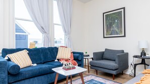 Ein Wohnzimmer mit einem blauen Sofa, einem grauen Sessel, einem kleinen Tisch mit Büchern und einem Bild an der Wand.