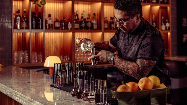 Ein Barkeeper bereitet Getränke an einer Bar zu, im Hintergrund ist eine gut sortierte Spirituosenregal zu sehen.