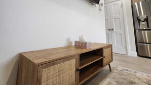 Una sala moderna con un televisor de pantalla plana montado en la pared, un mueble de madera para entretenimiento con una cesta tejida, y una zona de cocina con electrodomésticos de acero inoxidable.