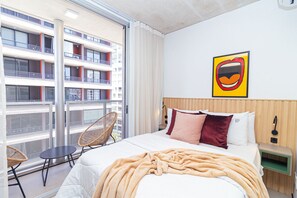 Habitación de hotel con una cama grande, vistas de un edificio desde un balcón, una silla y una obra de arte colgada en la pared.