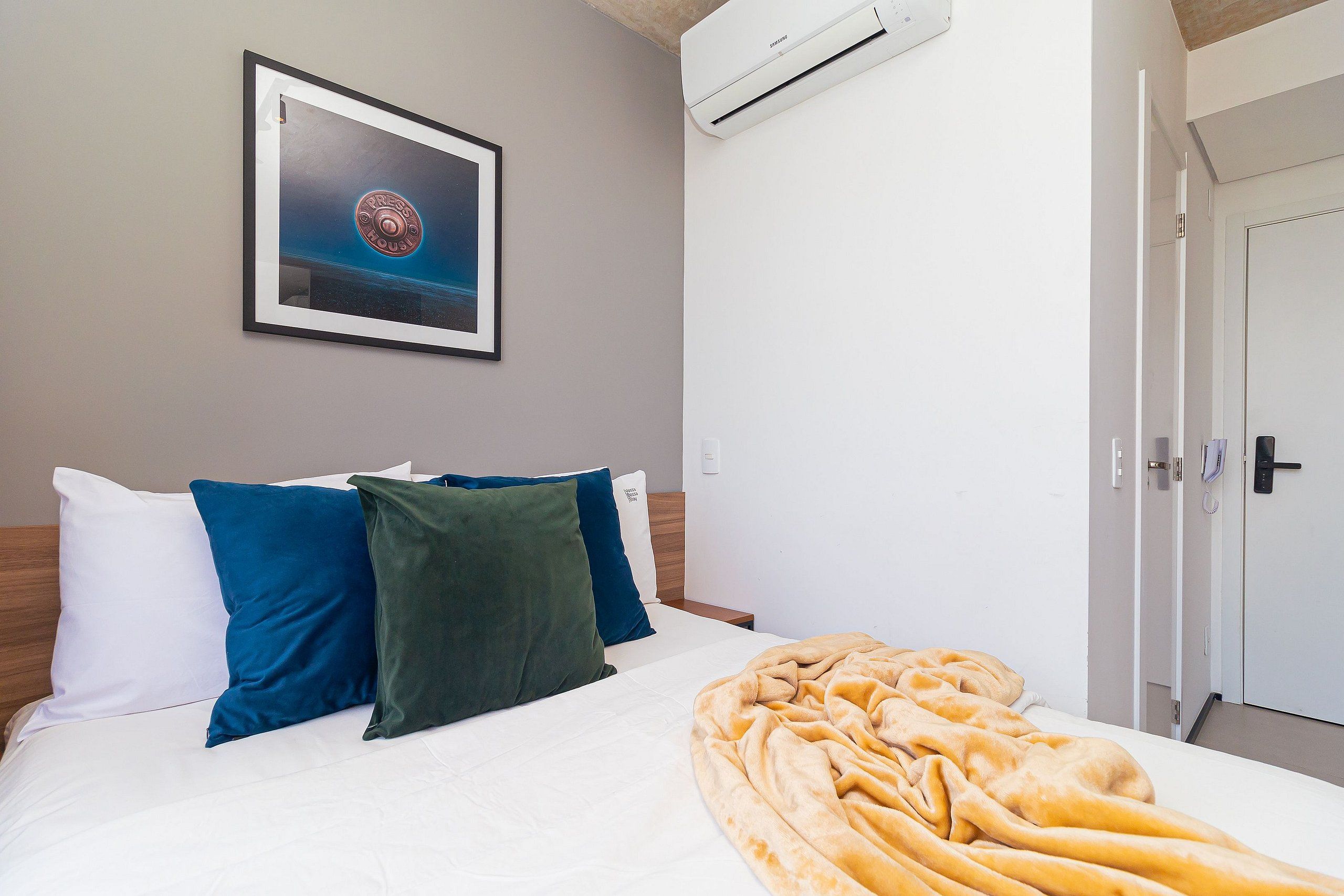 A neatly made bed with white and dark pillows, a framed picture on the wall, and an air conditioning unit above it.