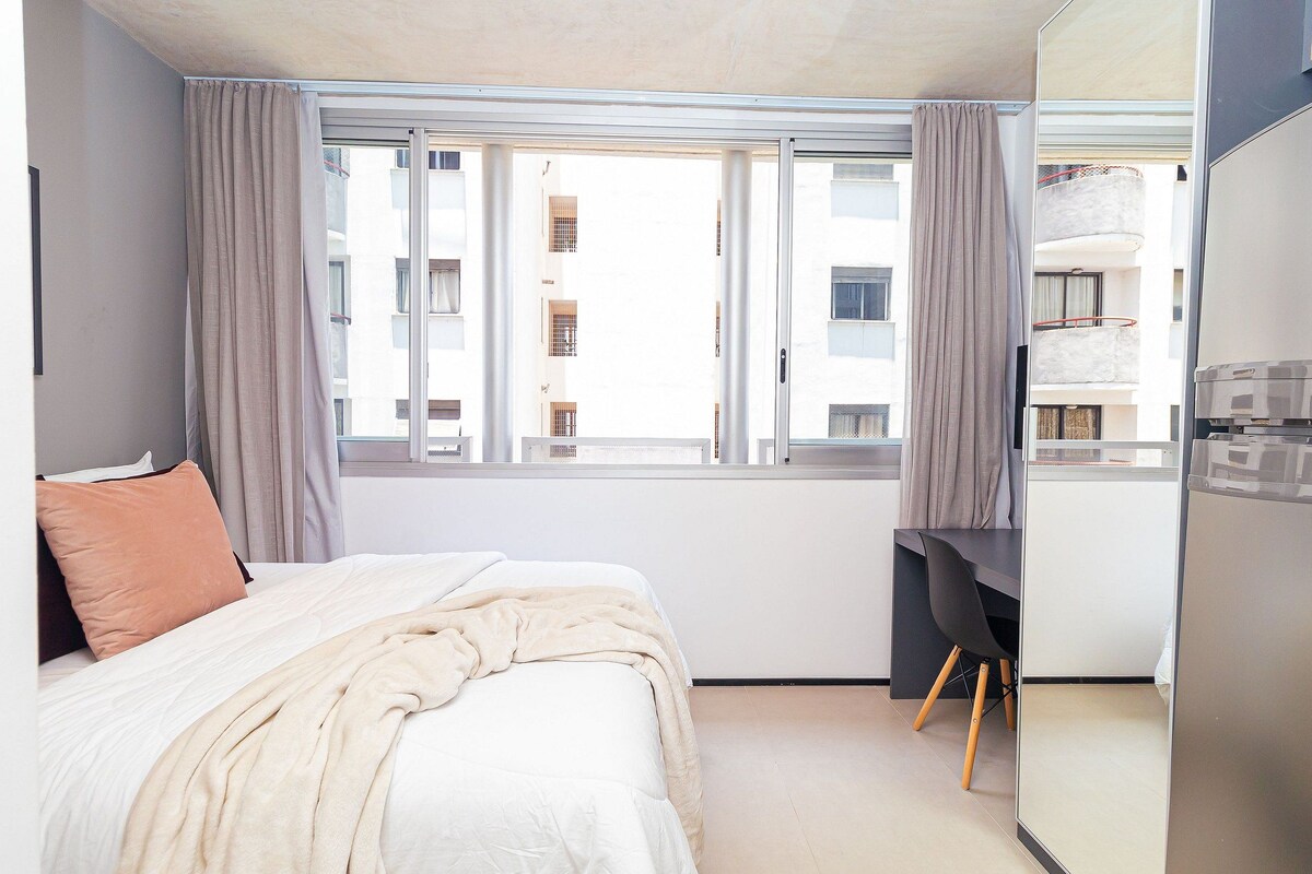 A bedroom with a bed, a desk, and a view of a building through a window.
