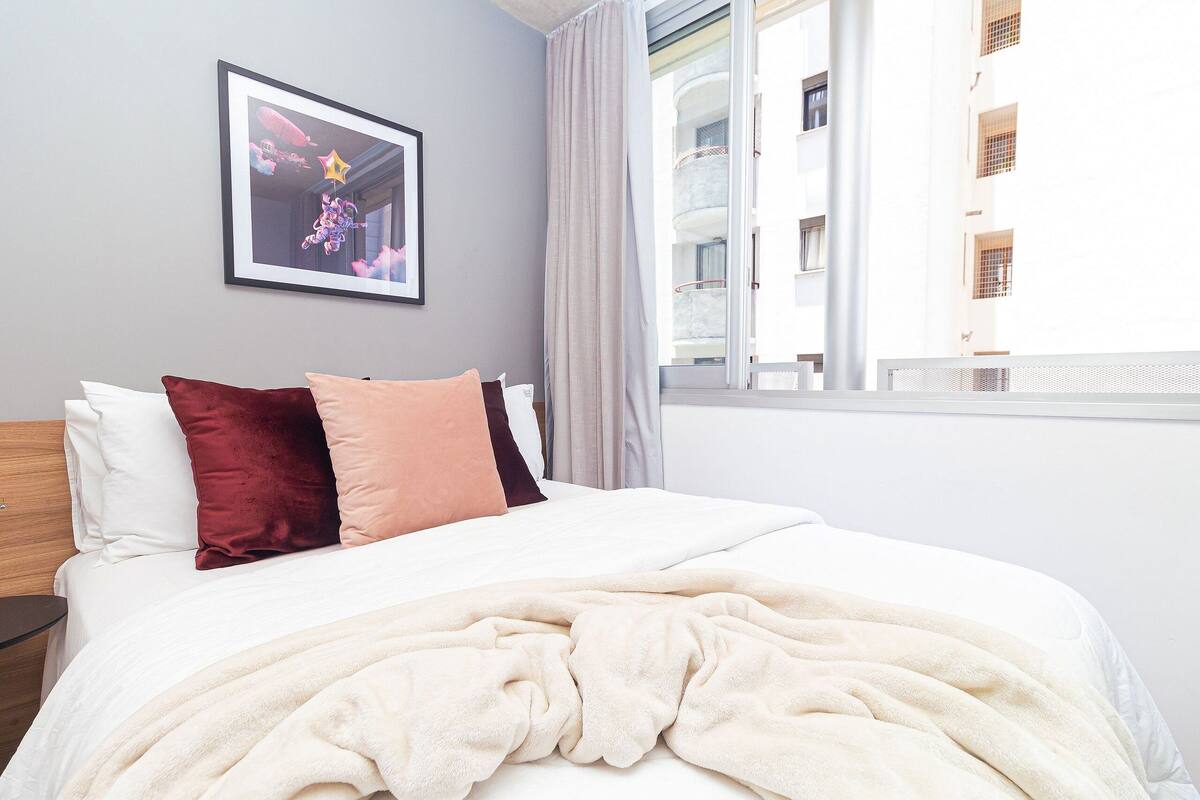 A neatly made bed with a white comforter and a beige blanket, a framed picture on the wall, and a view of a building through a window.