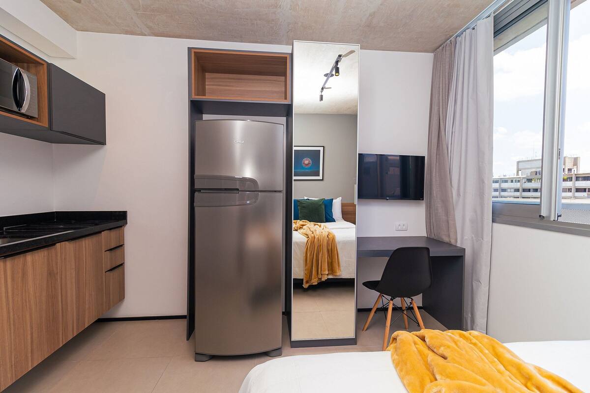 A compact hotel room with a kitchenette, a bed with a yellow blanket, a desk, and a TV.