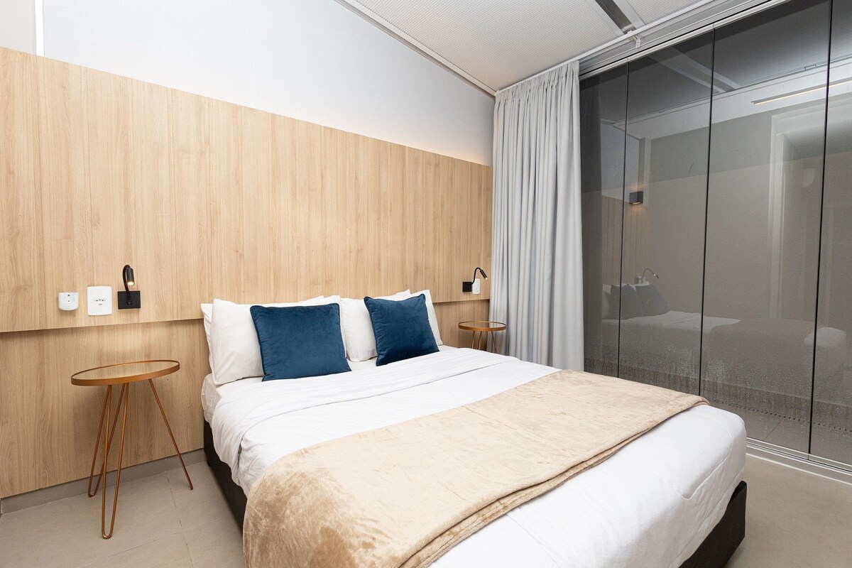 A modern hotel room with a large bed, a wooden accent wall, a small round table, and a sliding glass door leading to a balcony.