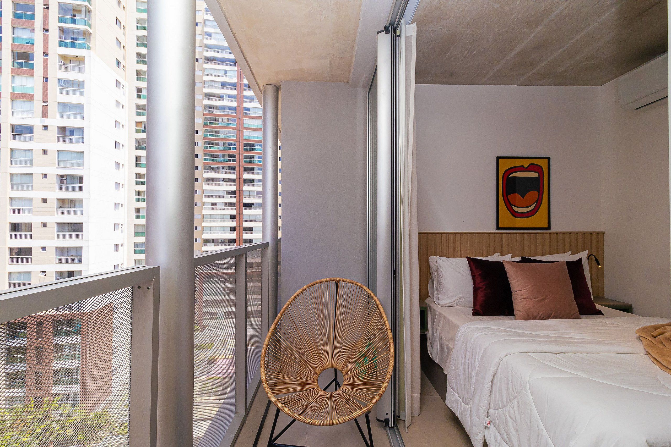 A balcony with a wicker chair, a bed with white bedding and two pillows, and a wall-mounted artwork.