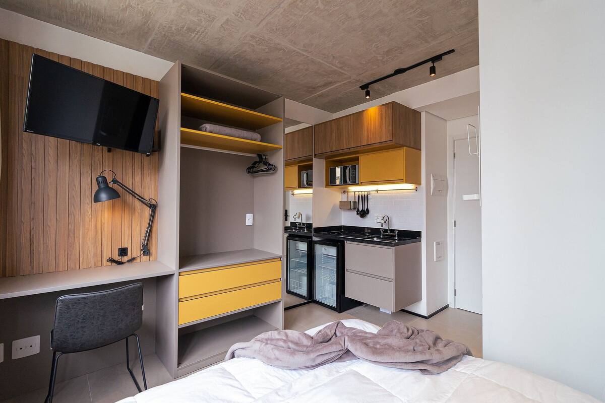 A modern hotel room with a bed, a desk, a chair, a TV, a kitchenette, and a bathroom.