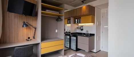 A compact hotel room with a bed, a desk, a chair, a TV mounted on the wall, a kitchenette, and a wardrobe.