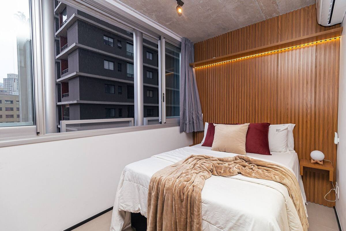 A modern hotel room with a large bed, wooden paneling on the walls, a window with a view of a building, and a bedside table with a lamp.