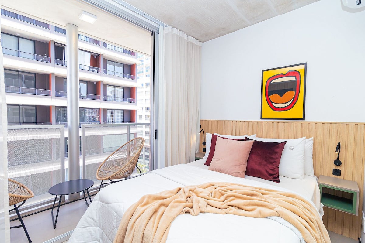 A hotel room with a large bed, a view of a building through a balcony, a chair, and a wall-mounted artwork.