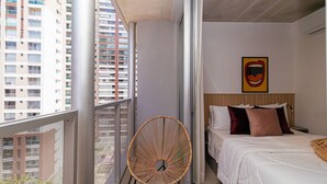 A balcony with a wicker chair, a bed with white bedding and two pillows, and a wall-mounted artwork. - Vossa Bossa Chez Frei (São Paulo)