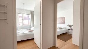 A modern hotel room with a wooden floor, a bed with white linens, a small bedside table with a vase, and a view of the outside through a window. - 2ndhomes Modern & Cozy 2BR Apt w Balcony (Helsinki)
