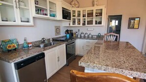 A kitchen with white cabinets, granite countertops, a dishwasher, and a dining area with a wooden chair.