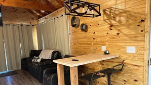 Un accogliente interno di una baita con una parete in legno, un divano in pelle nera, un tavolo in legno e due sgabelli neri.