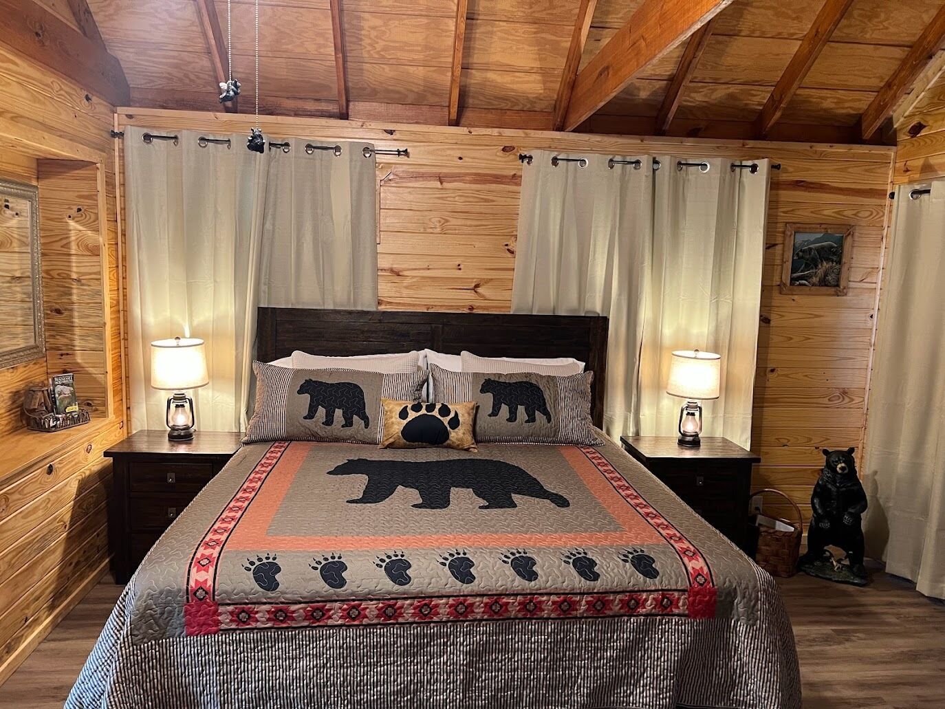 A cozy bedroom with a wooden ceiling, a bed with a bear-themed bedspread, two bedside lamps, and a small bear figurine on the nightstand.