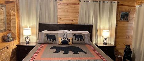 A cozy bedroom with a wooden ceiling, a bed with a bear-themed bedspread, two bedside lamps, and a small bear figurine on the nightstand.