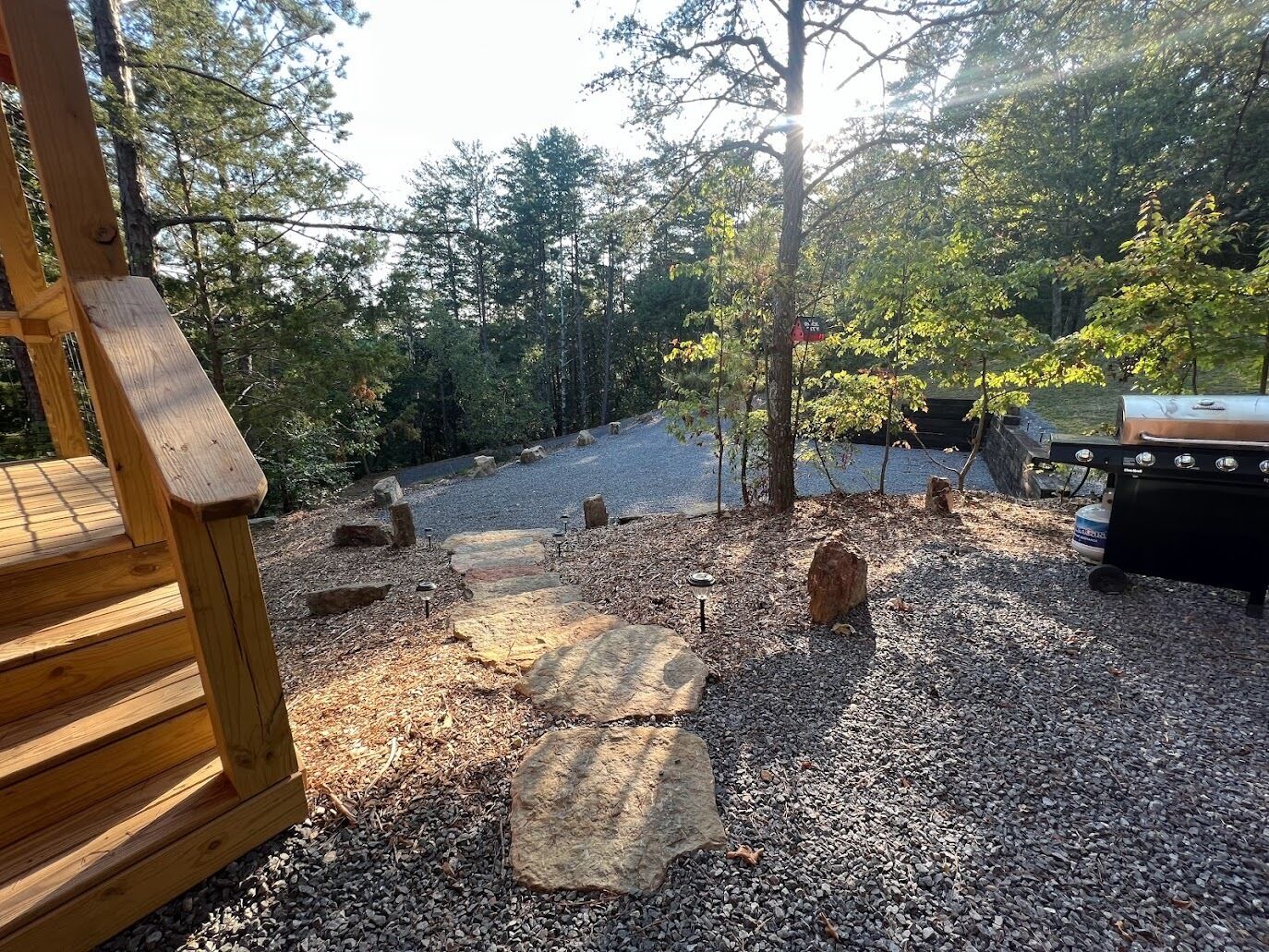 Eine Holzterrasse mit einem Steinweg, der zu einem Grillbereich inmitten von Bäumen führt.