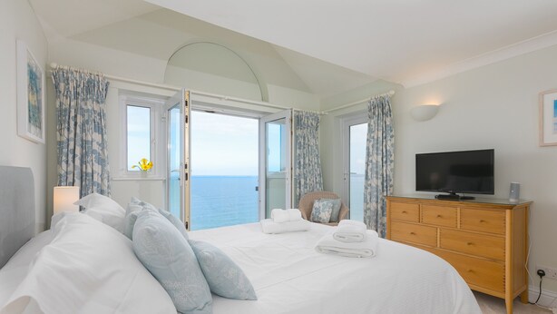 A bedroom with a large bed, a television, a dresser, and a view of the sea through open windows. - Wavetop (St Ives)