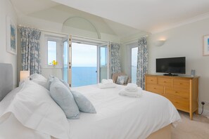A bedroom with a large bed, a television, a dresser, and a view of the sea through open windows.