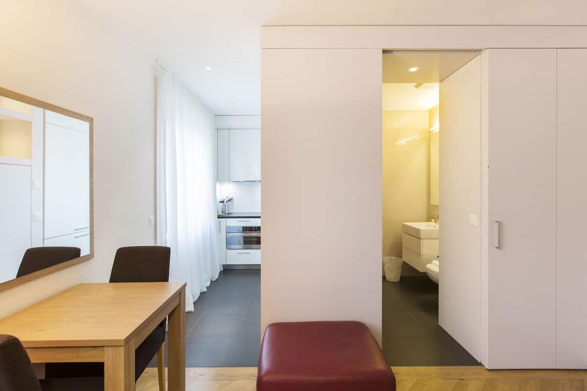 Une chambre d’hôtel moderne avec une table à manger en bois, un banc rouge et une vue sur la cuisine et la salle de bain.