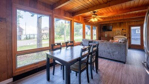 A wooden interior with a dining table and chairs, a kitchen area, and large windows offering a view of the outdoors.