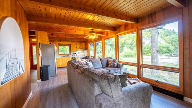 A cozy cabin interior with a large gray sectional sofa, wooden ceiling, and large windows offering a view of the outdoors.