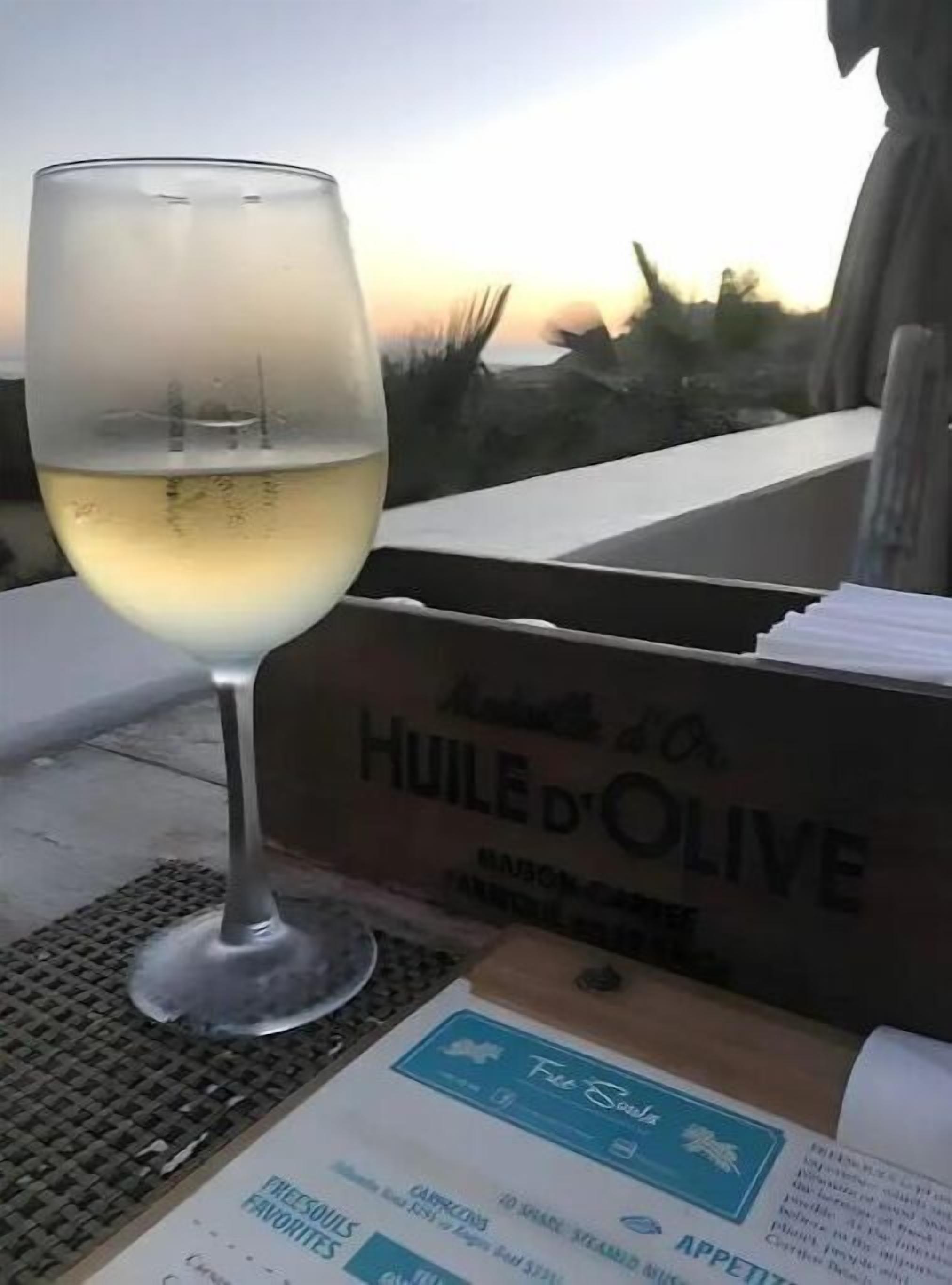Ein Glas Weißwein, eine Speisekarte und eine Schachtel mit der Aufschrift 'HUILE D'OLIVE' auf einer Terrasse.