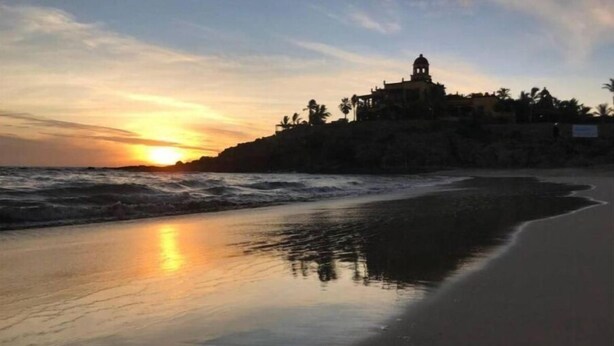 A beach with a building on a cliff, waves crashing onto the shore, and a sunset in the background. - 1A Casa Un Amor, Cerritos Beach Condo (El Pescadero)