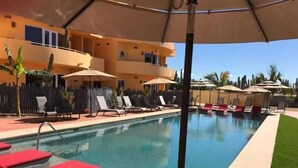 A swimming pool with lounge chairs and umbrellas, surrounded by a fence and a building with balconies. - 1A Casa Un Amor, Cerritos Beach Condo (El Pescadero)