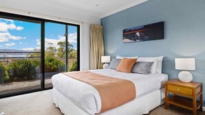 A bedroom with a large bed, two bedside tables, a window with a view of greenery and a distant landscape, and a painting on the wall. - Stockyards 1 with uninterrupted mountain and lake views (Jindabyne)