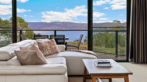 A balcony with a white sofa, a wooden table, and a view of a lake and mountains. - Stockyards 1 with uninterrupted mountain and lake views (Jindabyne)