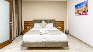 A neatly made bed with white linens and a grey headboard, a small trash bin beside the bed, and a framed picture of a building on the wall. - Luxurious, spacious fully equipped home in Swieqi (Is-Swieqi)