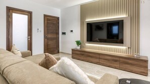 A modern living room with a large flat-screen TV mounted on a wooden wall, a beige sofa, and a wooden cabinet below. - Luxurious, spacious fully equipped home in Swieqi (Is-Swieqi)
