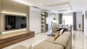 A modern living room with a large TV, built-in shelves, a dining area, and a sofa. - Luxurious, spacious fully equipped home in Swieqi (Is-Swieqi)