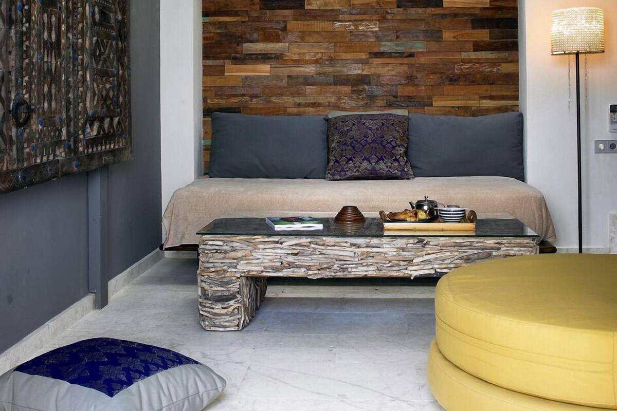 Een moderne slaapkamer met een schuine plafond, een houten hoofdbord, een bed met grijze beddengoed, een nachtkastje met boeken en een dienblad, een vloerlamp en een groot abstract schilderij.