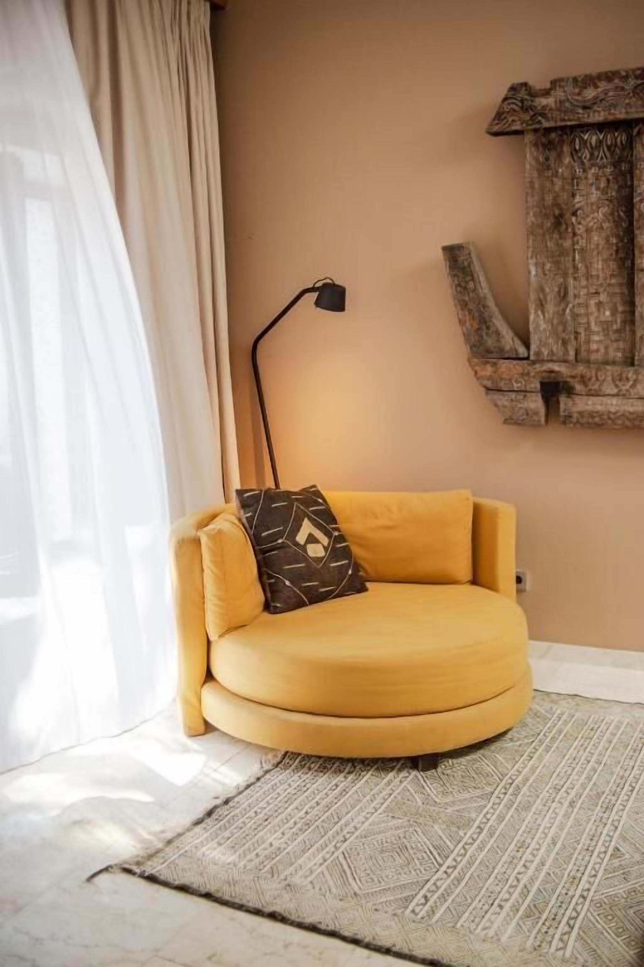 Un sillón amarillo con un cojín de estampado geométrico, una lámpara de pie y un adorno de pared de madera rústica en una habitación con pared de color claro y alfombra estampada.