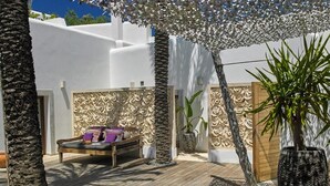 Een houten terras met een bank en kussens, omgeven door palmbomen en een decoratieve muur.