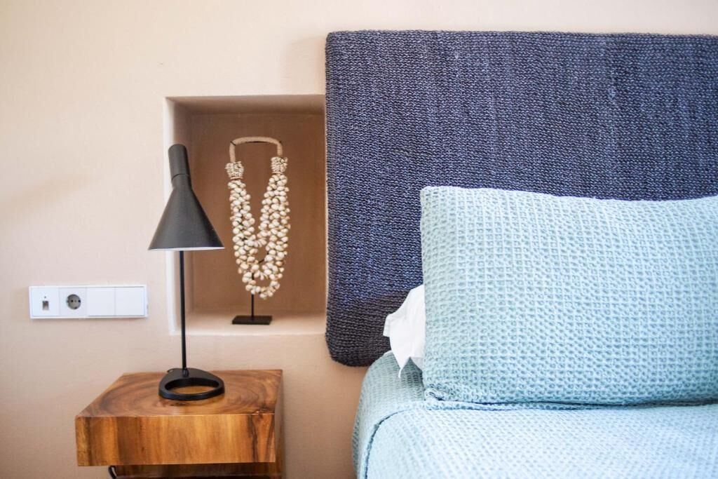 Quarto bem arrumado com mesa de cabeceira de madeira, abajur preto, colar de contas decorativo, almofada azul texturizada e cabeceira azul escura.
