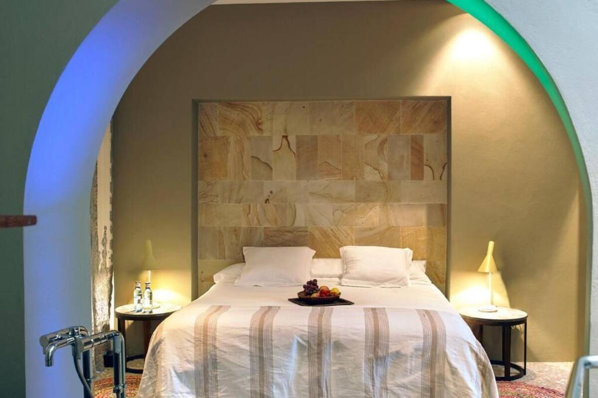 Hotelkamer met een groot bed, een stenen hoofdeinde, twee nachtkastjes met lampen en een schaal fruit op het bed.