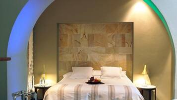 Habitación de hotel con una cama grande, cabecera de piedra, dos mesitas de noche con lámparas y una bandeja de frutas sobre la cama.